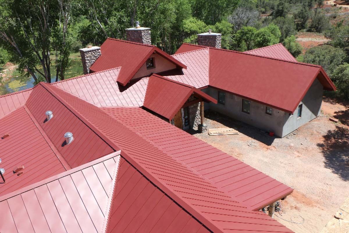 Red metal roof on a multi-sectioned house, chimneys, and green trees in the background.