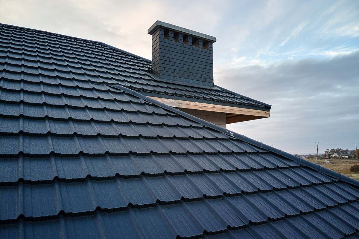 Dark blue metal roof tiles and a chimney under a cloudy sky.