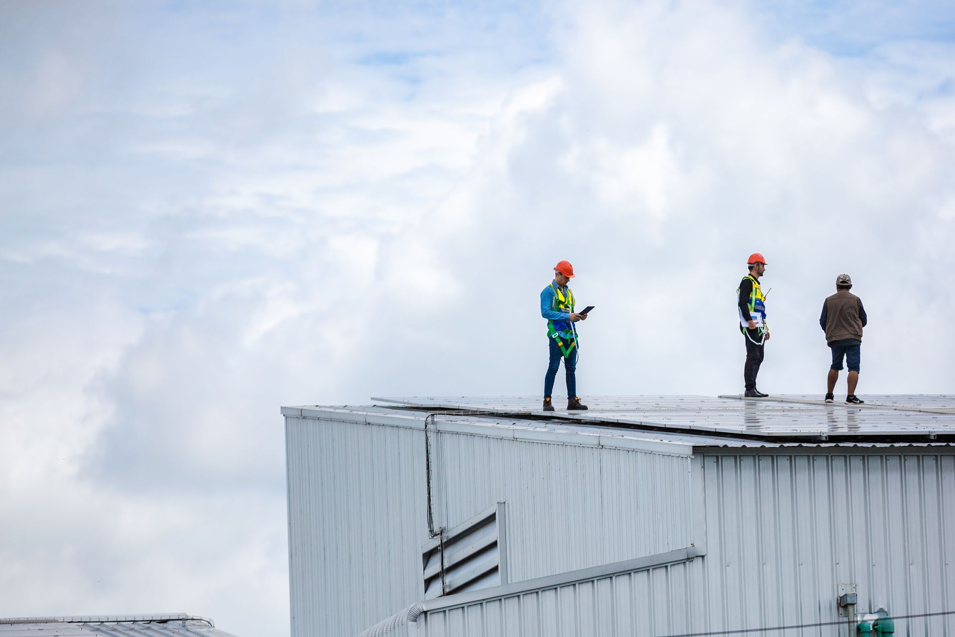Three workers on a rooftop, wearing hard hats and safety harnesses, against a cloudy sky.