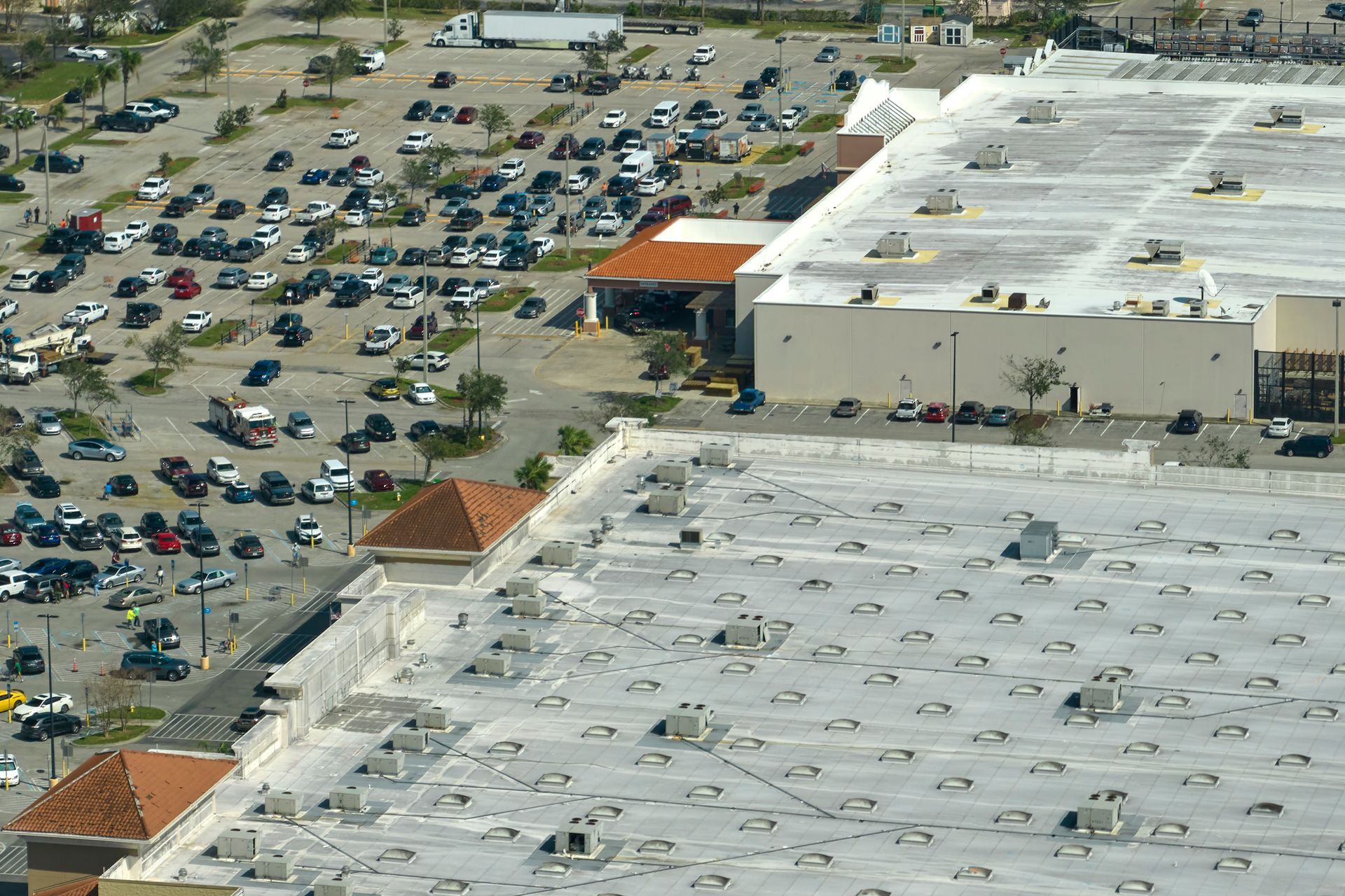 Aerial view of a shopping center and full parking lot. White and beige buildings with many cars parked outside.
