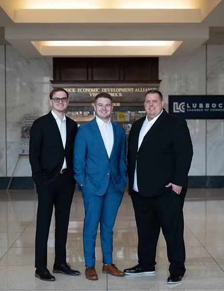 Three men in suits standing in a building lobby. The man in the middle wears a blue suit, the others black. They are smiling.