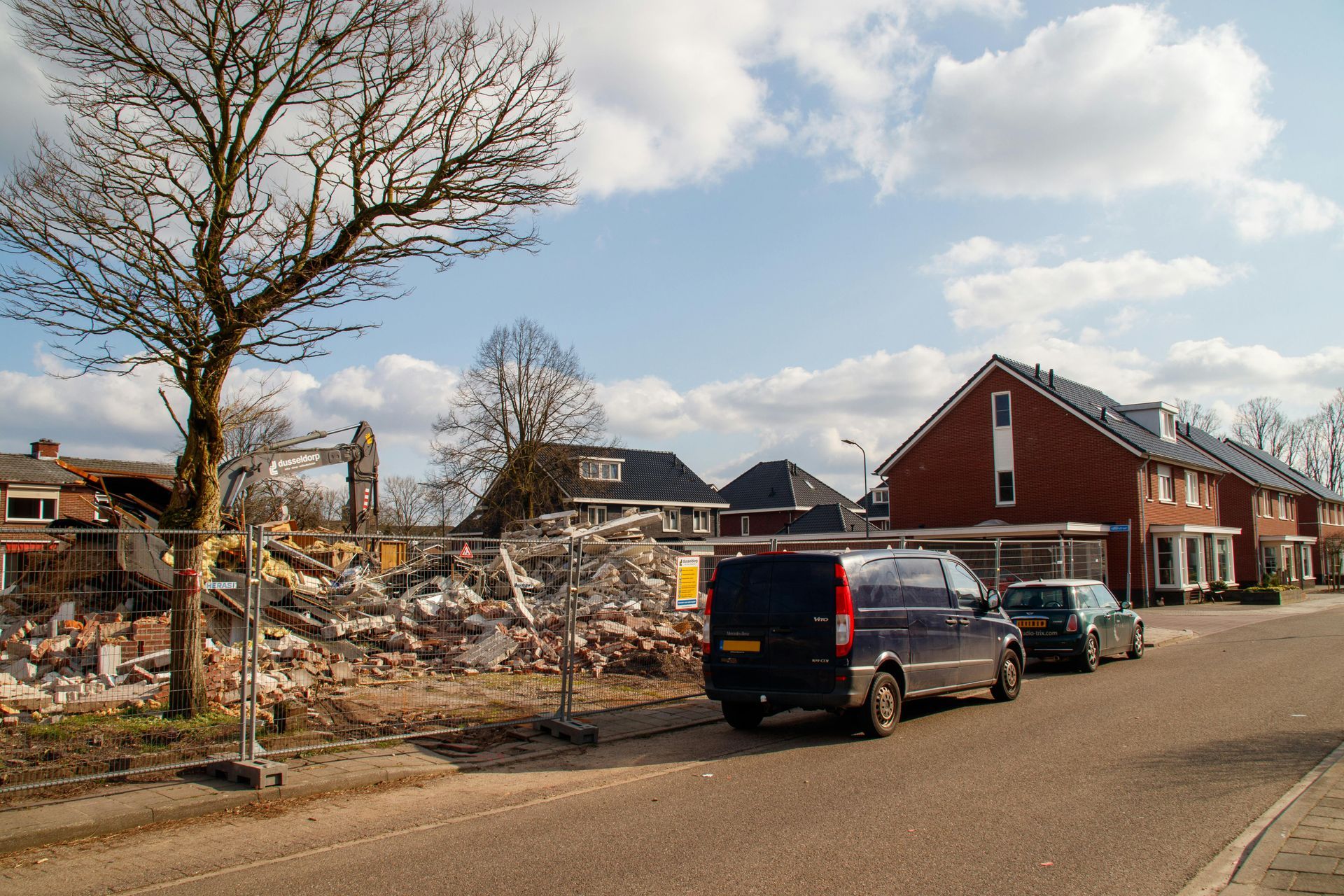 Demolition site with rubble behind a fence, next to a street with parked cars and houses under a blue sky.