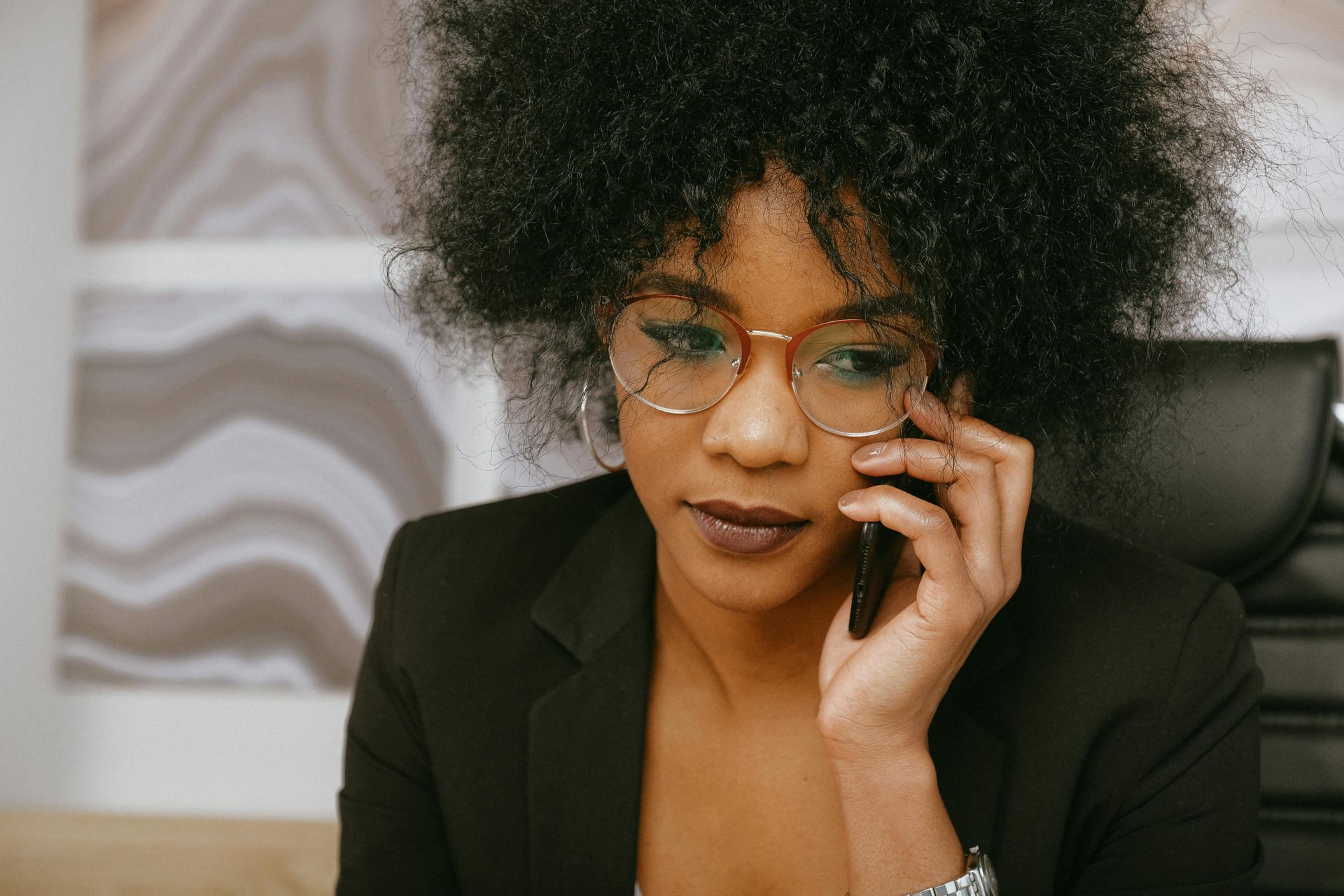 Woman with dark, curly hair, wearing glasses and a blazer, talking on a phone in an office setting.