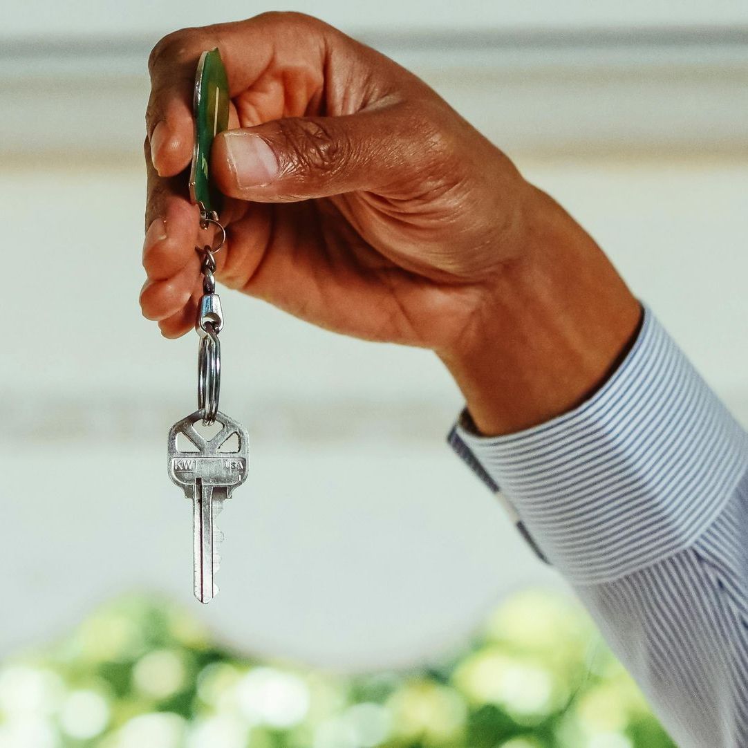 Hand holding up a set of keys, possibly a home key, in front of a blue-striped shirt and a blurred background.