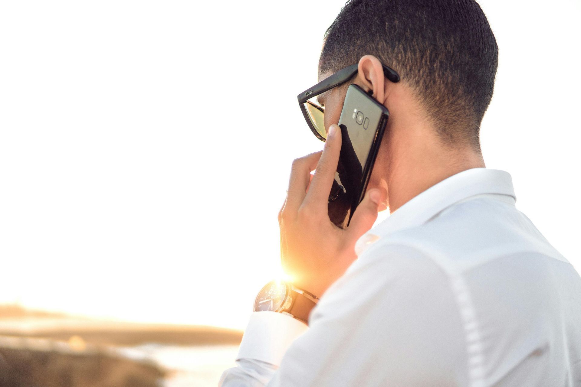 A man in a white shirt and sunglasses holds a phone to his ear, silhouetted against a bright sunset.