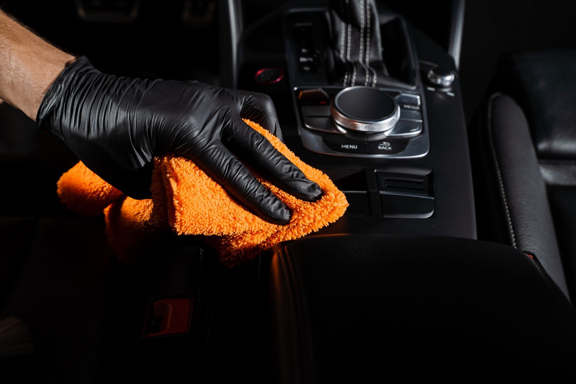 Gloved hand wiping a car's black interior with an orange microfiber cloth.