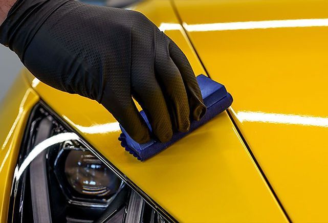 Gloved hand applying ceramic coating to yellow car paint with a blue applicator.