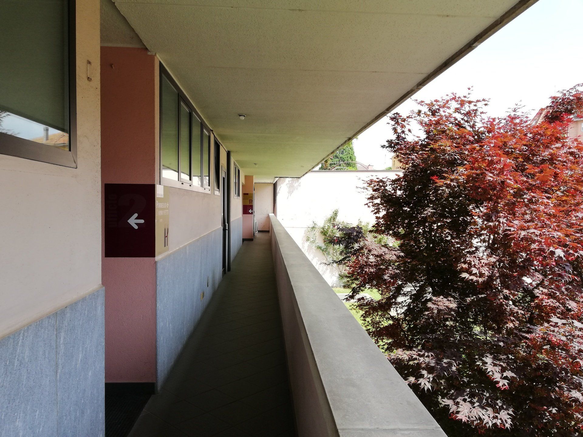 Passerella esterna con un albero rosso sulla destra e un muro rosa e blu sulla sinistra. Un cartello con freccia indica la direzione verso sinistra.