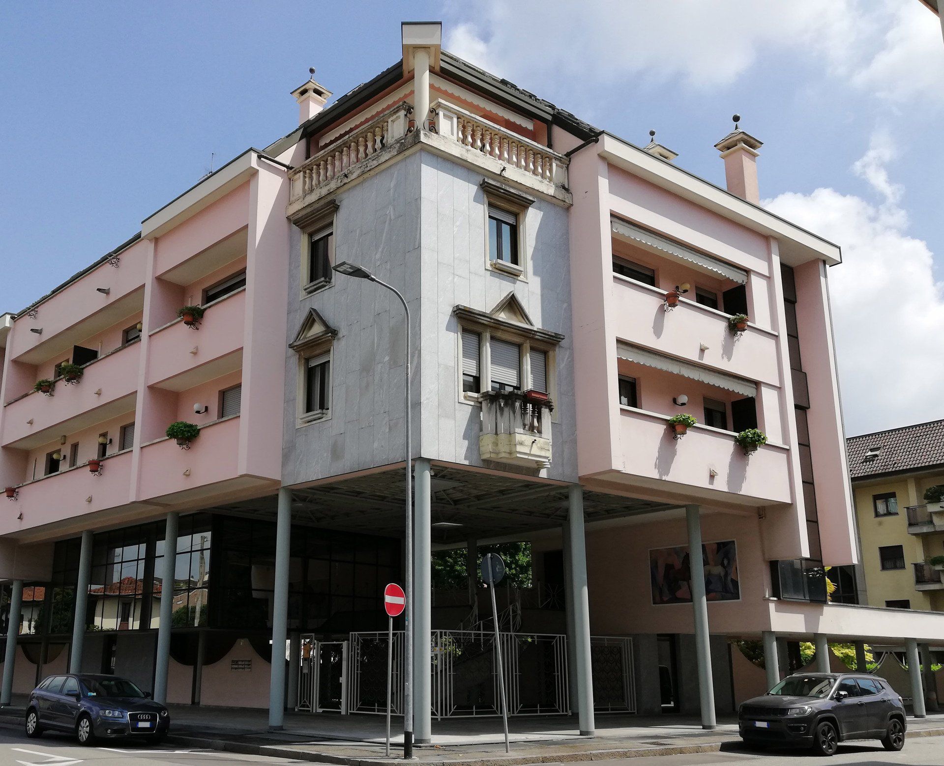 Edificio rosa e bianco a più piani con balconi e un primo piano rialzato sostenuto da pilastri. Auto parcheggiate di fronte.