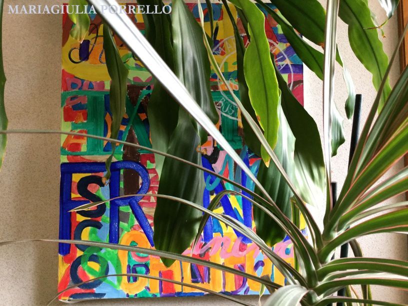 Opera d'arte astratta colorata con testo, parzialmente nascosta da piante verdi.