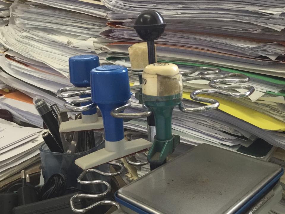 Francobolli in un contenitore su una scrivania ingombra di documenti, una penna nera e una piccola bilancia.