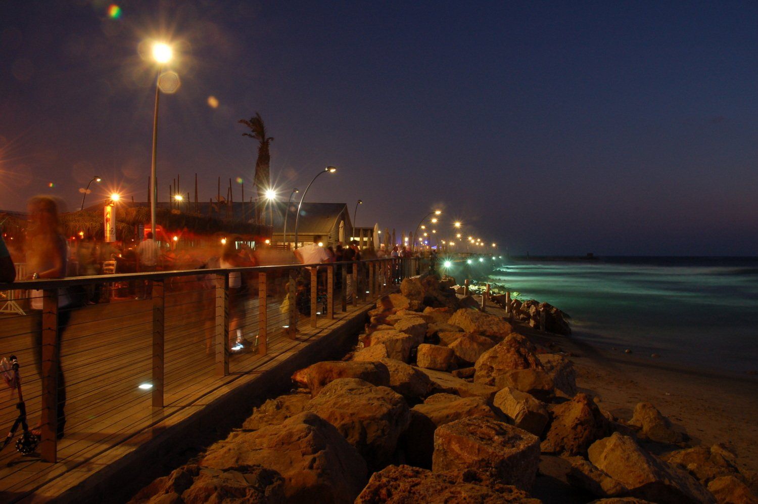 נוף לילי של מזח על חוף הים עם אורות המשתקפים על מים וסלעים לאורך החוף.