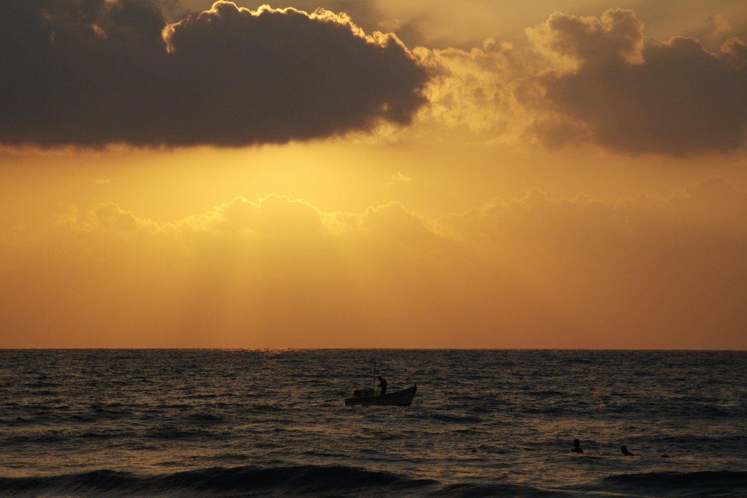 שקיעה מעל האוקיינוס עם צללית של סירה קטנה, אור זהוב פורץ מבעד לעננים.