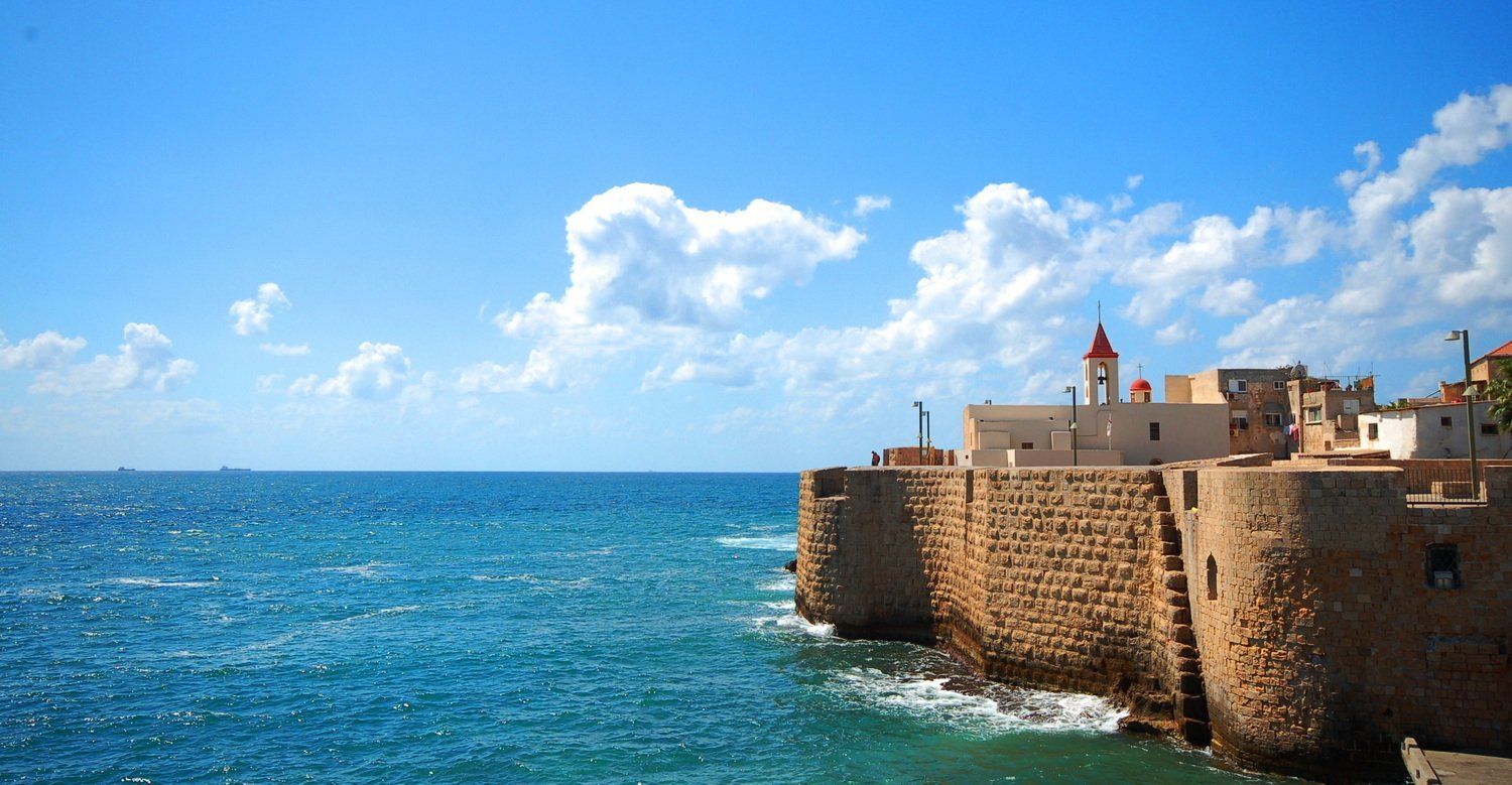 חומות האבן של עכו, ישראל, פוגשות את הים הטורקיז תחת שמיים כחולים. בניינים לבנים עם כנסייה אדומה.