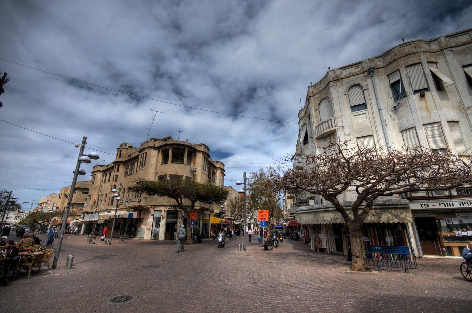 סצנת רחוב ביפו, ישראל, עם בניינים ישנים, חנויות, אנשים הולכים ושמיים מעוננים.