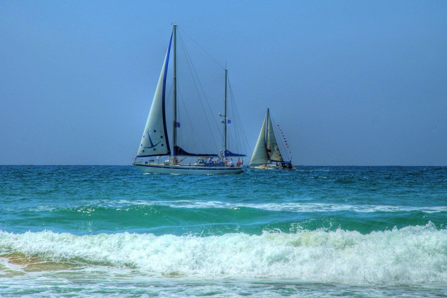 שתי סירות מפרש על מים כחולים, עם מפרשים לבנים על רקע שמיים בהירים. גלים מתנפצים בחזית.