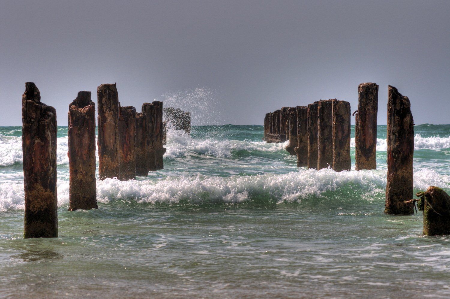 גלי אוקיינוס מתנפצים על עמודי רציף מעץ בלויים; שמיים מעוננים.