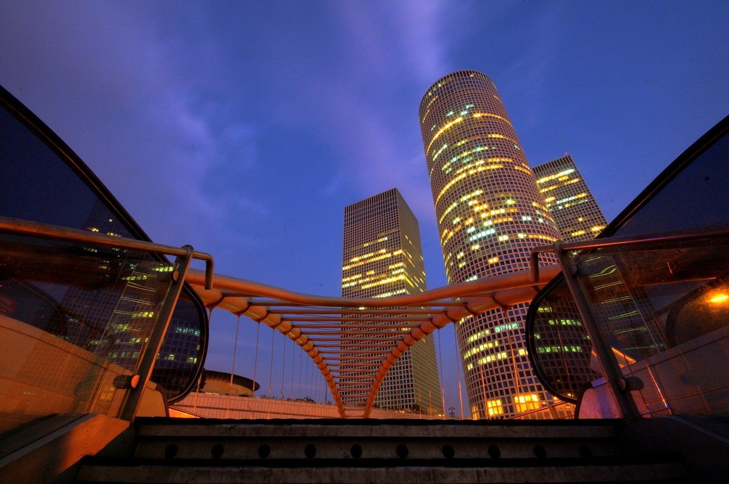 קו רקיע של עיר מודרנית עם רדת החשיכה. גורדי שחקים מוארים מתנשאים מעל גשר ומדרגות זהובים, מתחת לשמיים כחולים.