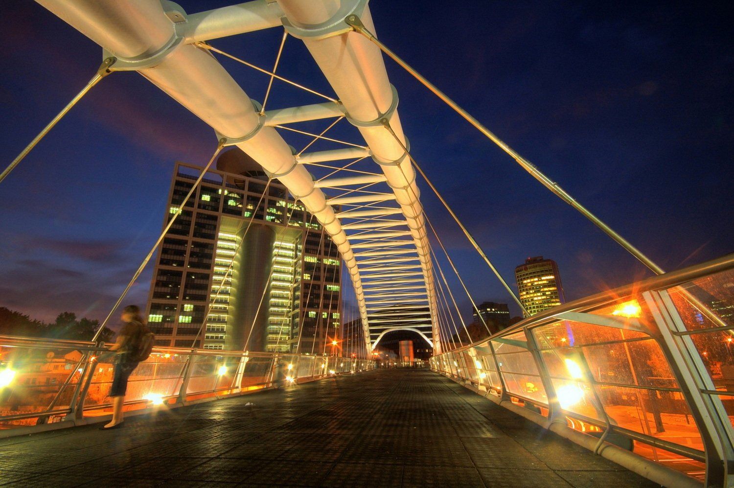 גשר להולכי רגל בלילה עם בנייני עיר ברקע. אדם עומד משמאל. אורות בהירים.