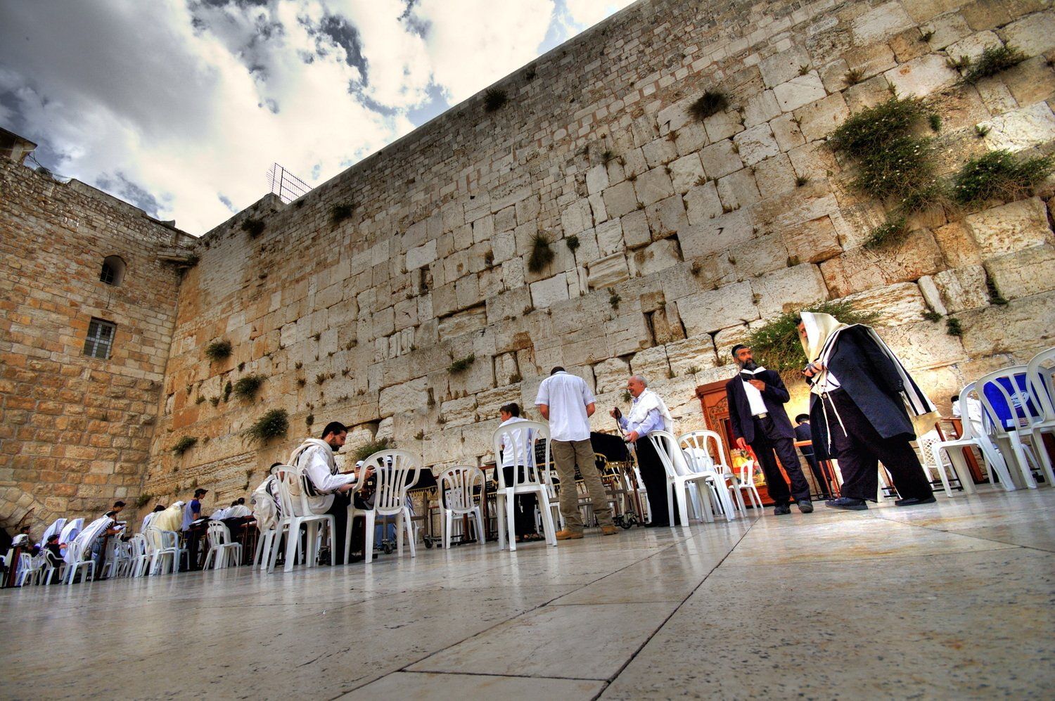 אנשים מתפללים בכותל המערבי בירושלים. קיר אבן גדול עם אנשים בטליתות.