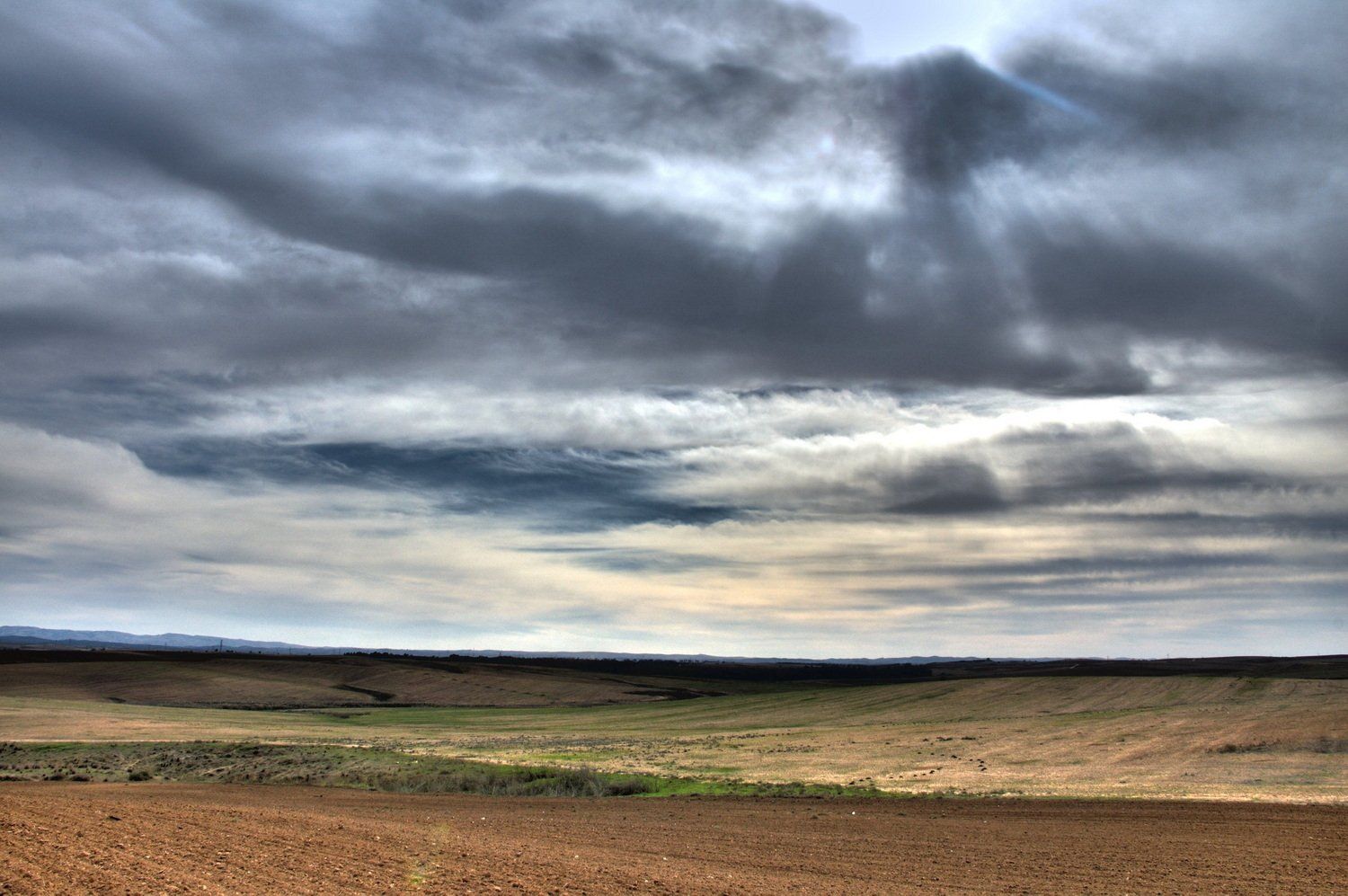 שמיים מעוננים מעל שדה רחב ידיים, חום וירוק. אור שמש מציץ מבעד לעננים כהים.