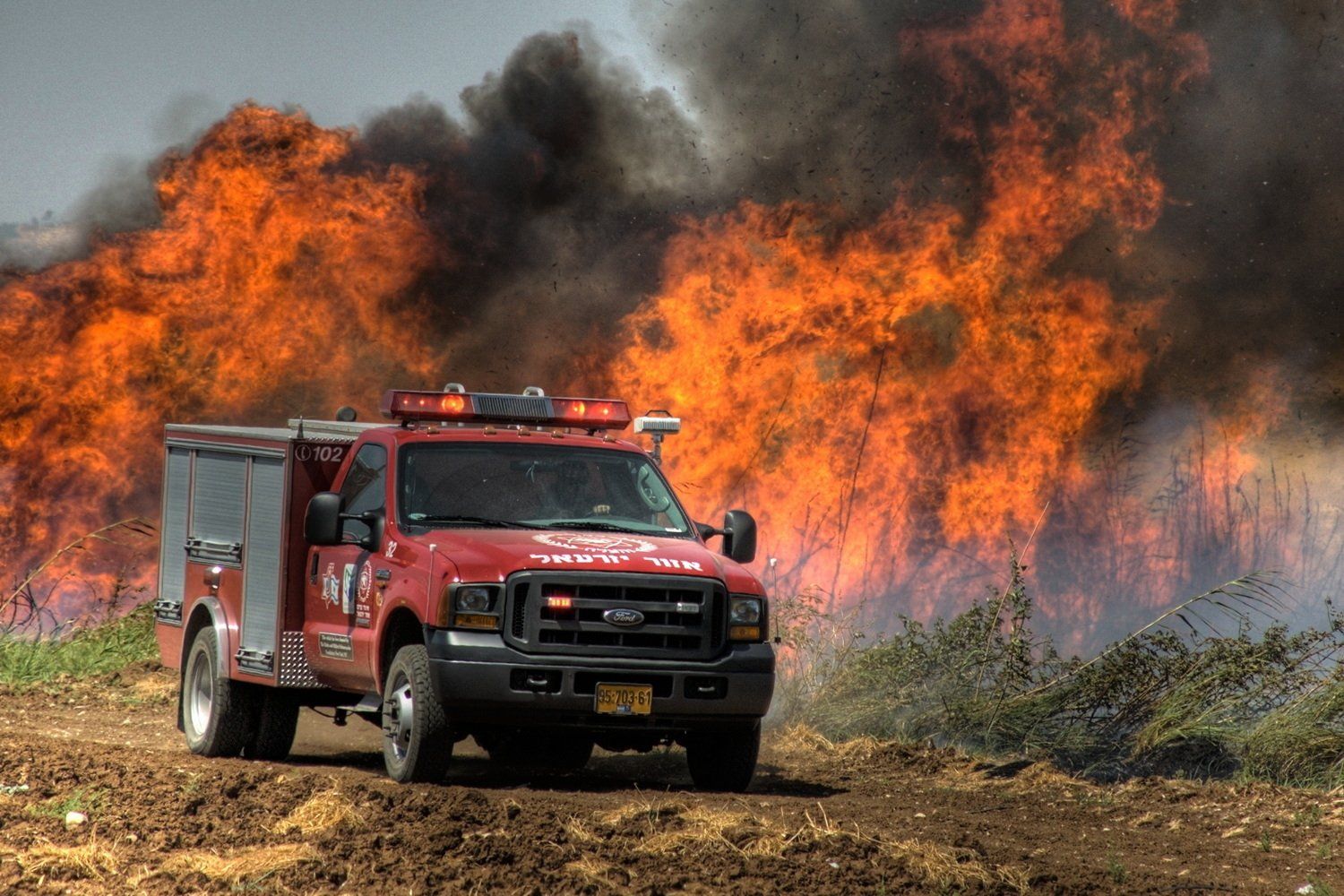 משאית כיבוי אש עומדת בפני שריפת יער גדולה עם להבות כתומות ועשן שחור.
