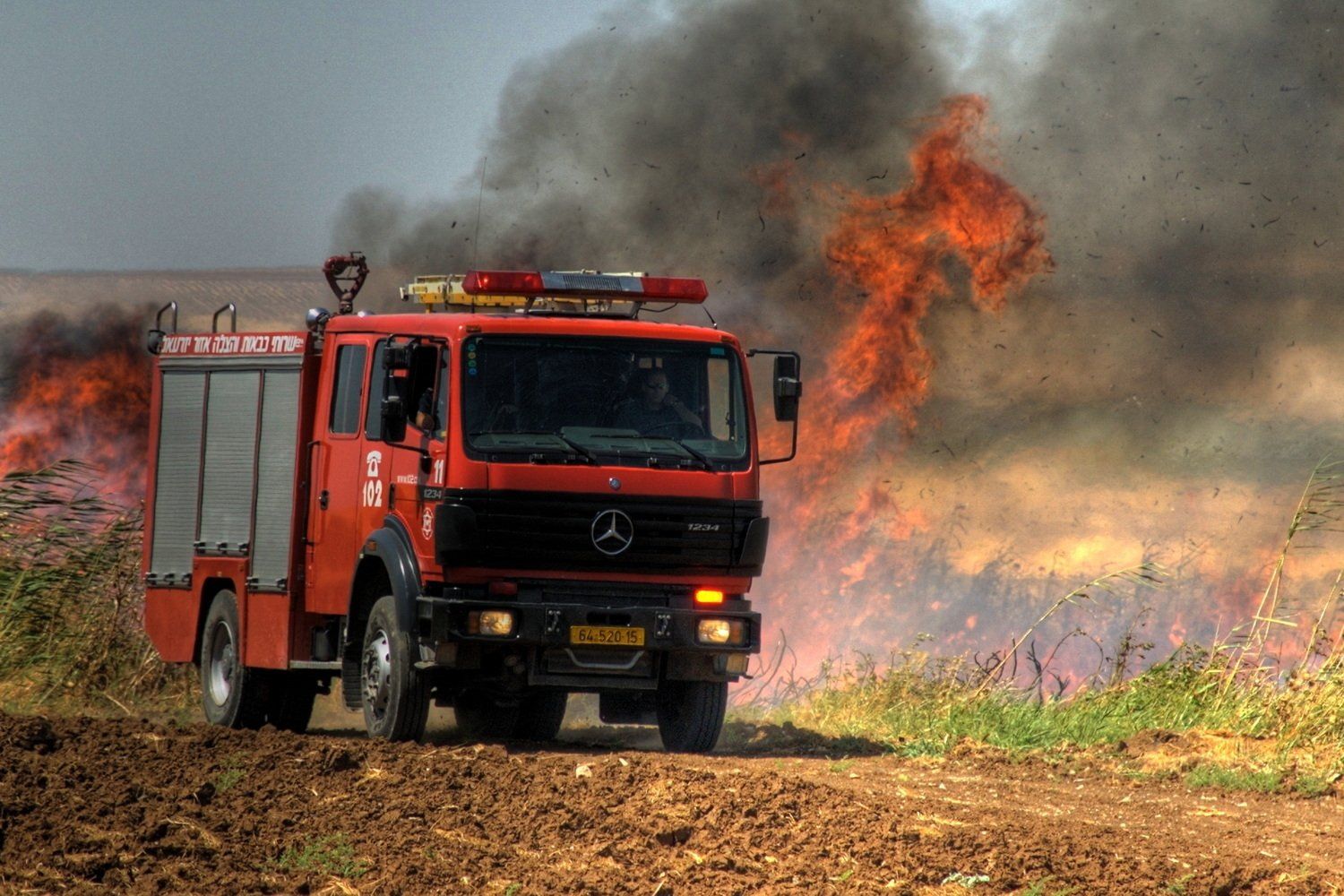 משאית כיבוי אדומה נאבקת בשריפה, כשלהבות ועשן מיתמרים בשדה.