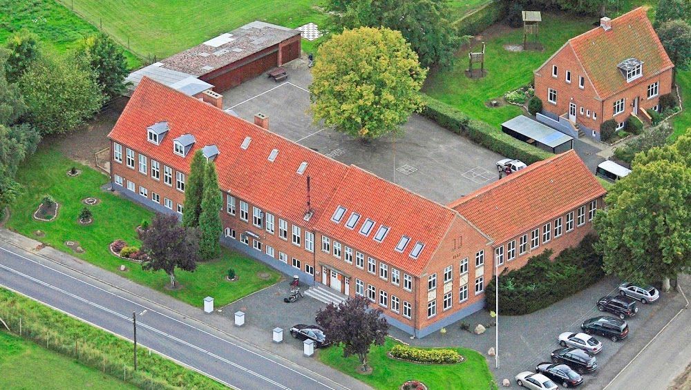 Luftfoto af en skolebygning af mursten med rødt tag, en gårdsplads, grønne omgivelser og parkerede biler.