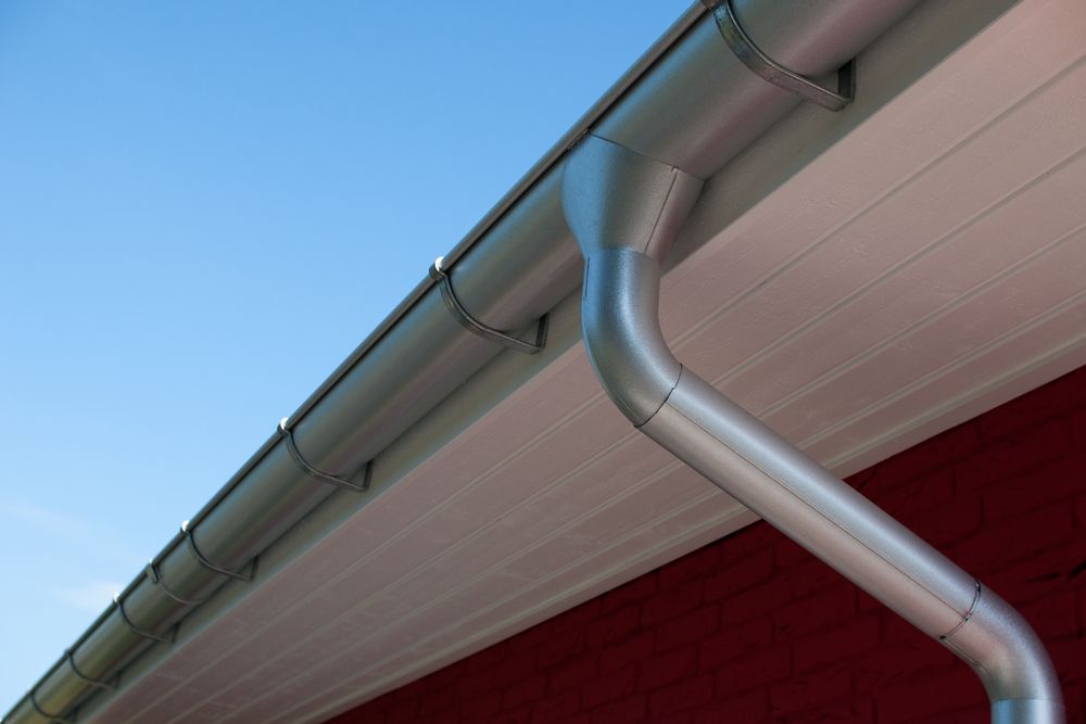 A silver metal gutter system and downspout attached to the white eaves of a building with a red brick wall  — B & A Plumbing & Gas In Wauchope, NSW