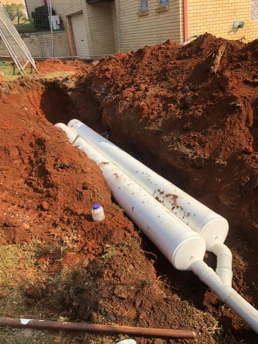 A trench in reddish soil contains two white PVC pipes connected by a Y-junction, with a house visible in the background — B & A Plumbing & Gas In Wauchope, NSW
