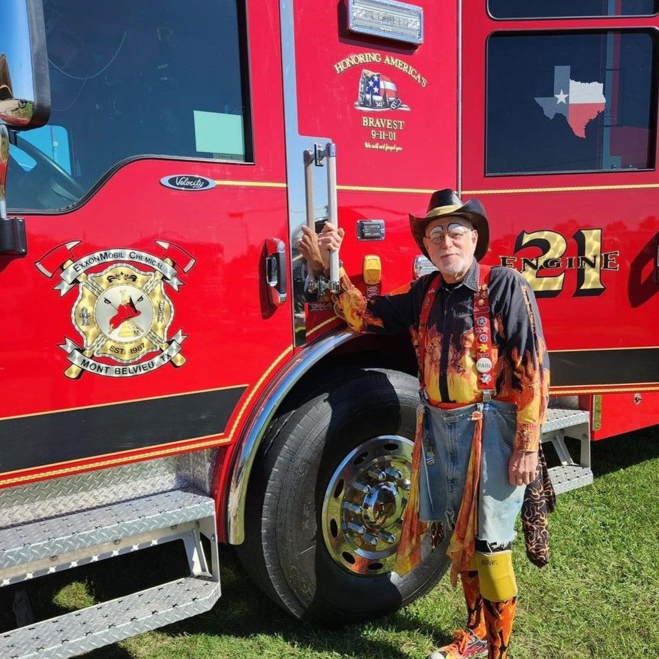 Man standing beside a bright red fire truck, wearing a cowboy hat and colorful clothes