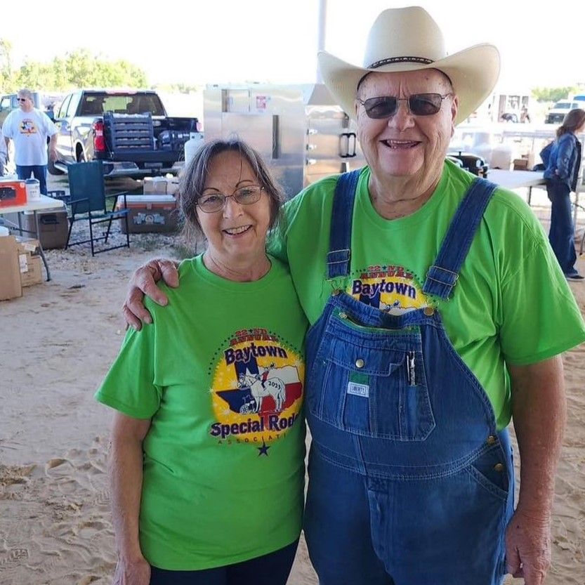 Two smiling people in green shirts at a fair, one in cowboy hat and overalls, with booths in the background