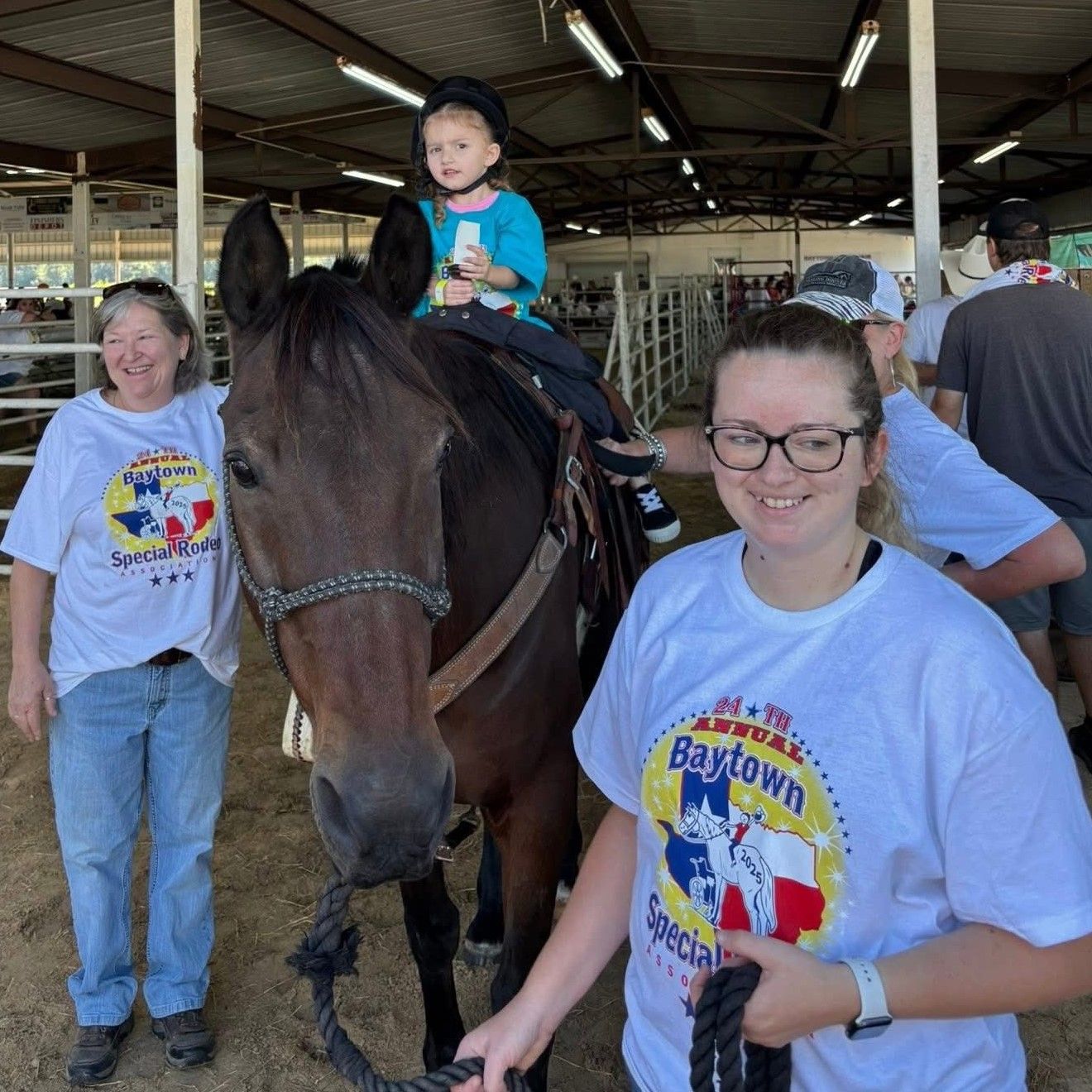 Child riding a horse at an indoor stable, smiling volunteers nearby in white T-shirts.