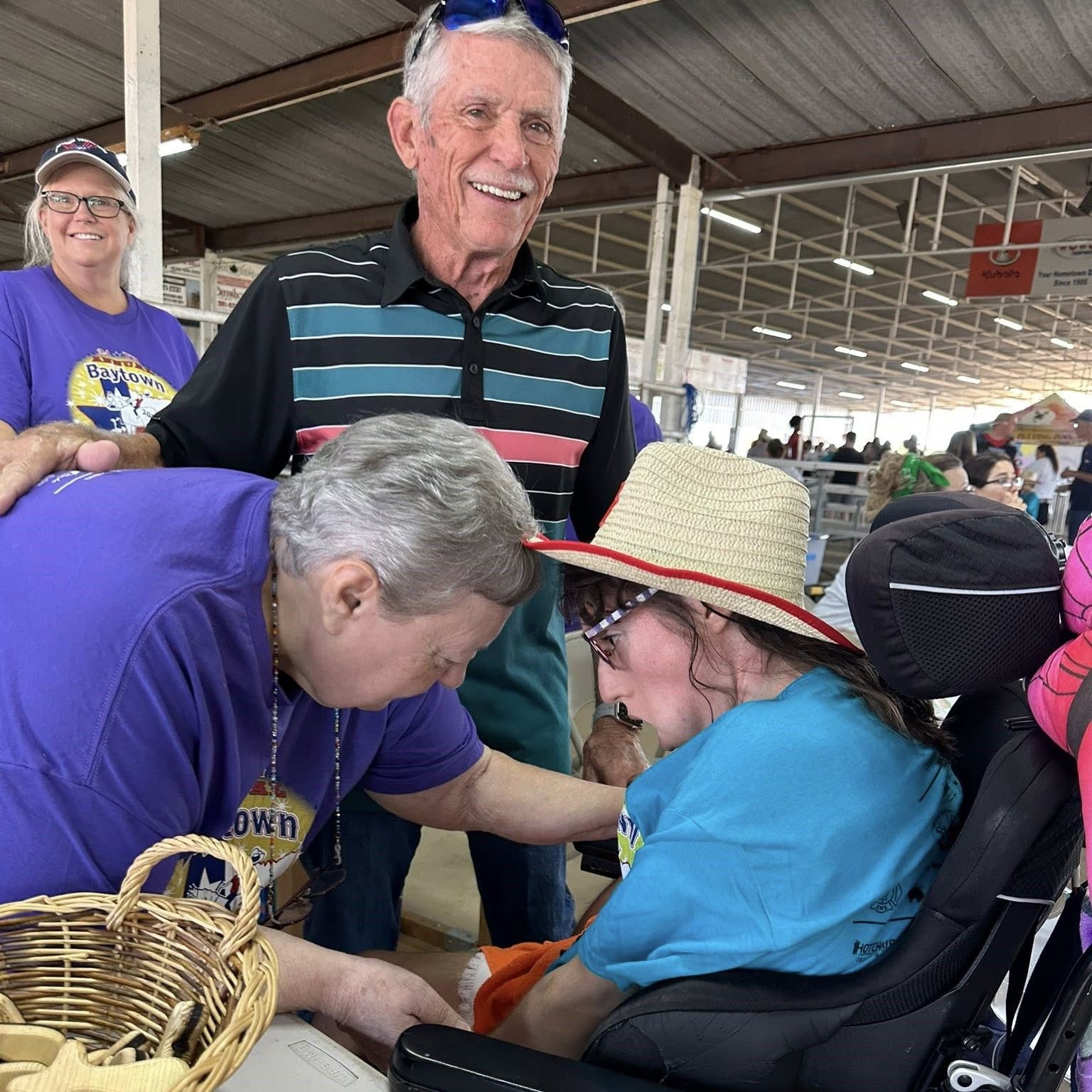 People at a fair help a seated person in a wheelchair under a pavilion.