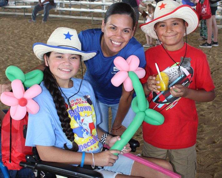 Three smiling people in cowboy hats holding balloon flowers at an outdoor event