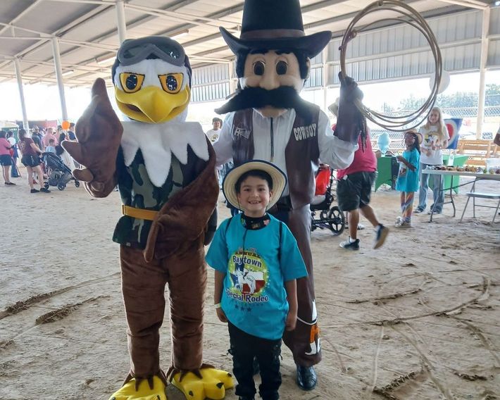 Child posing with two eagle mascots at an indoor event hall, one mascot waving.