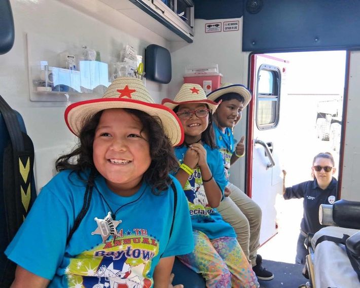 Smiling children in straw hats inside a van, posing by the open door while an adult waves outside.