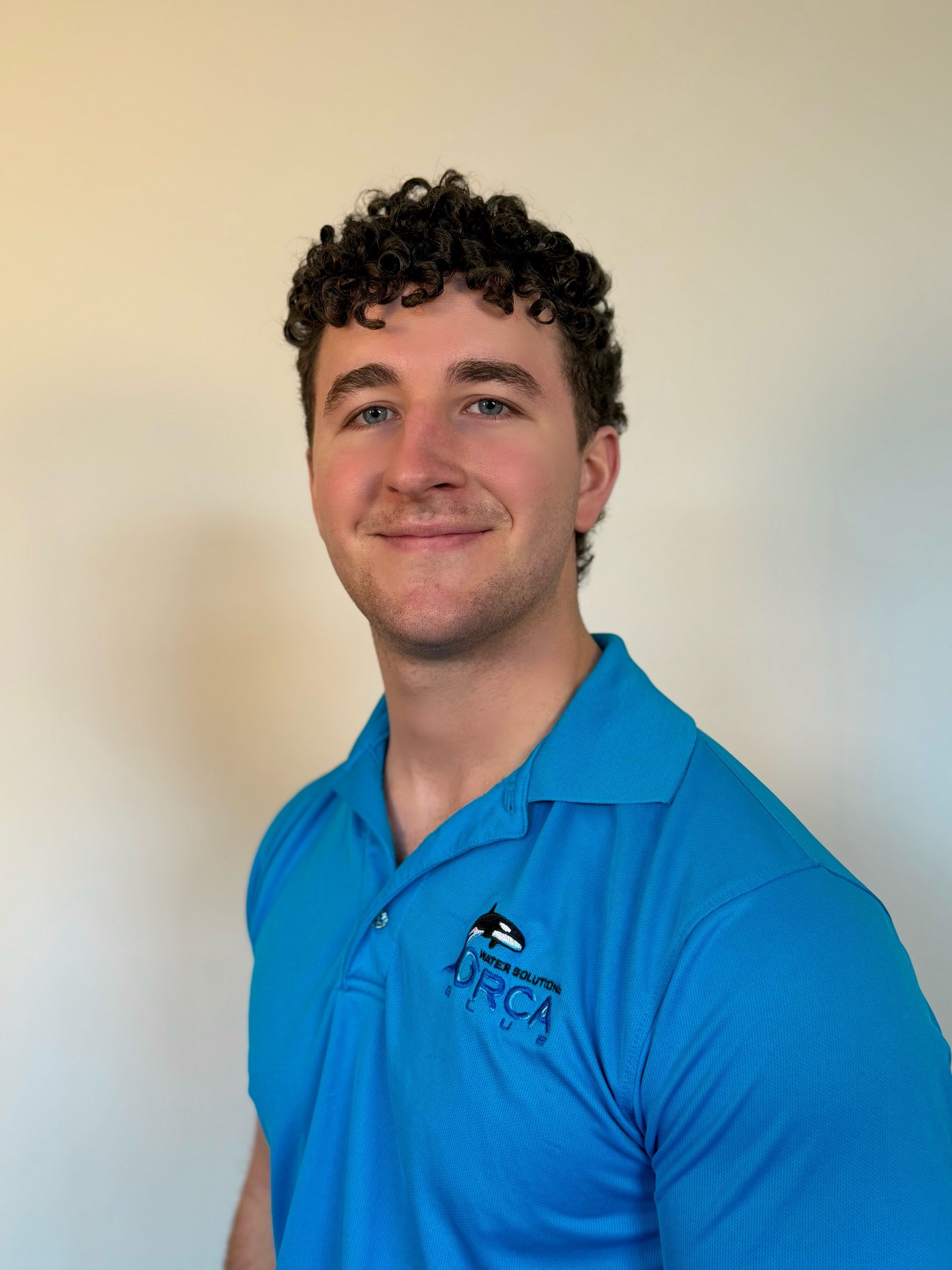 A person with curly hair smiling while wearing a bright blue polo shirt with an orca logo against a white background.