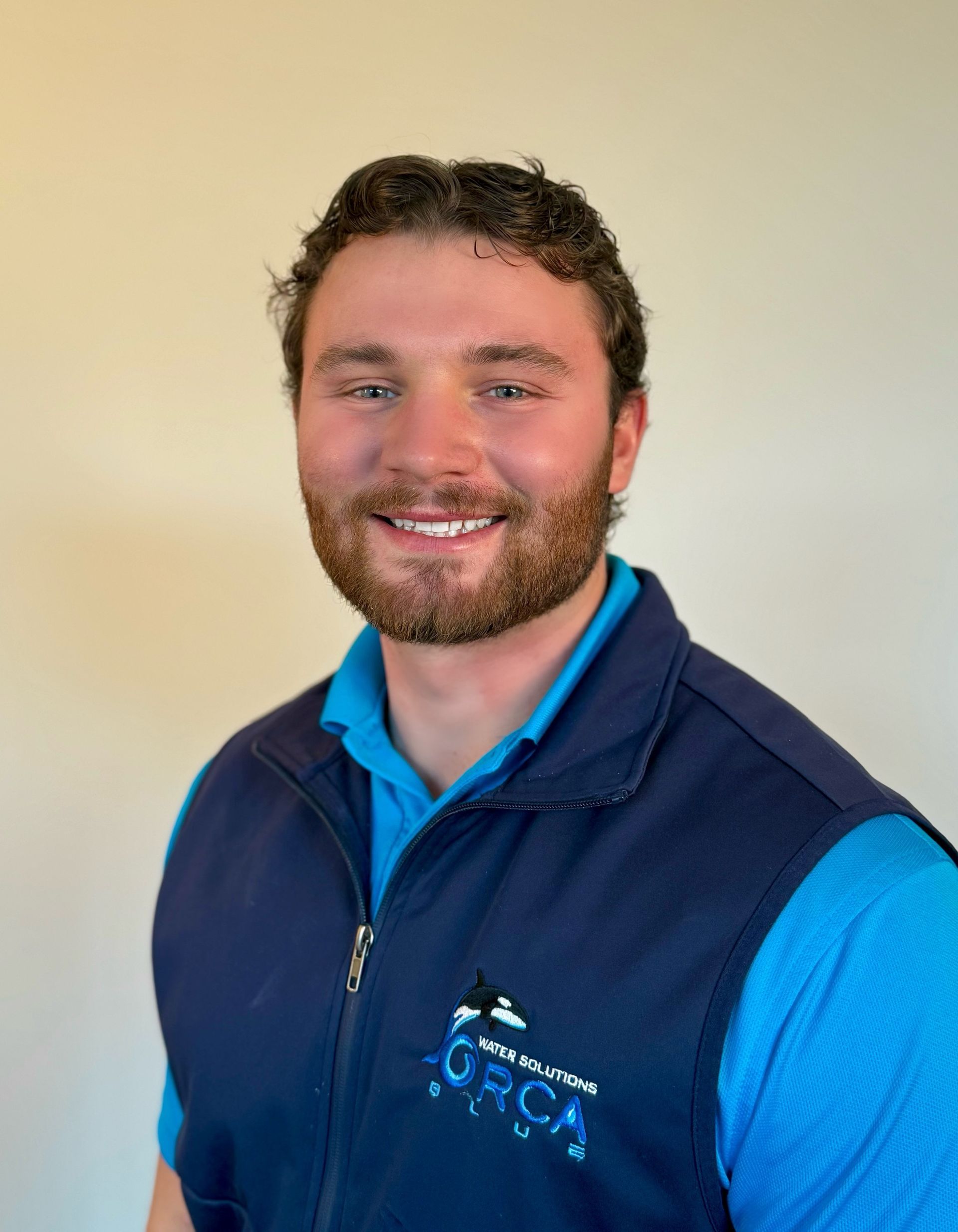 A smiling person with brown hair and a beard, wearing a bright blue collared shirt and a dark navy vest with a logo.