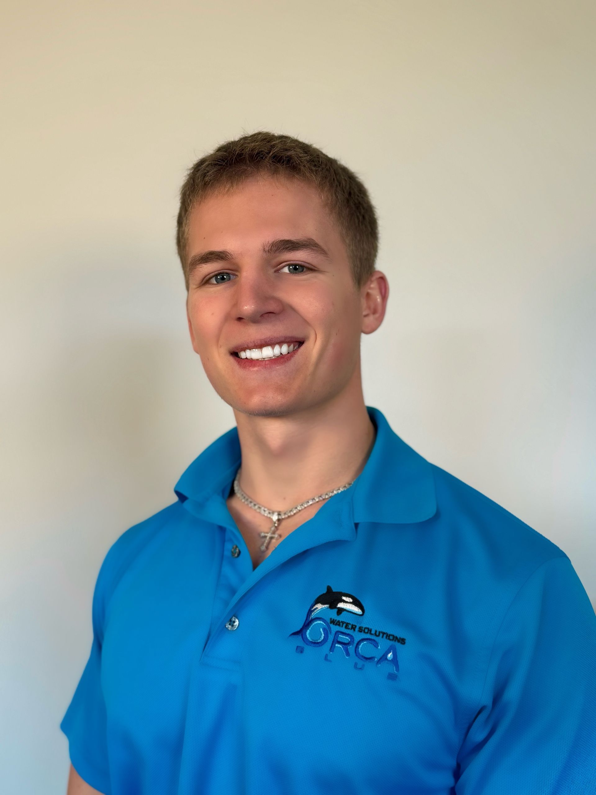 A person with short brown hair smiles while wearing a bright blue polo shirt featuring an orca logo against a plain wall.