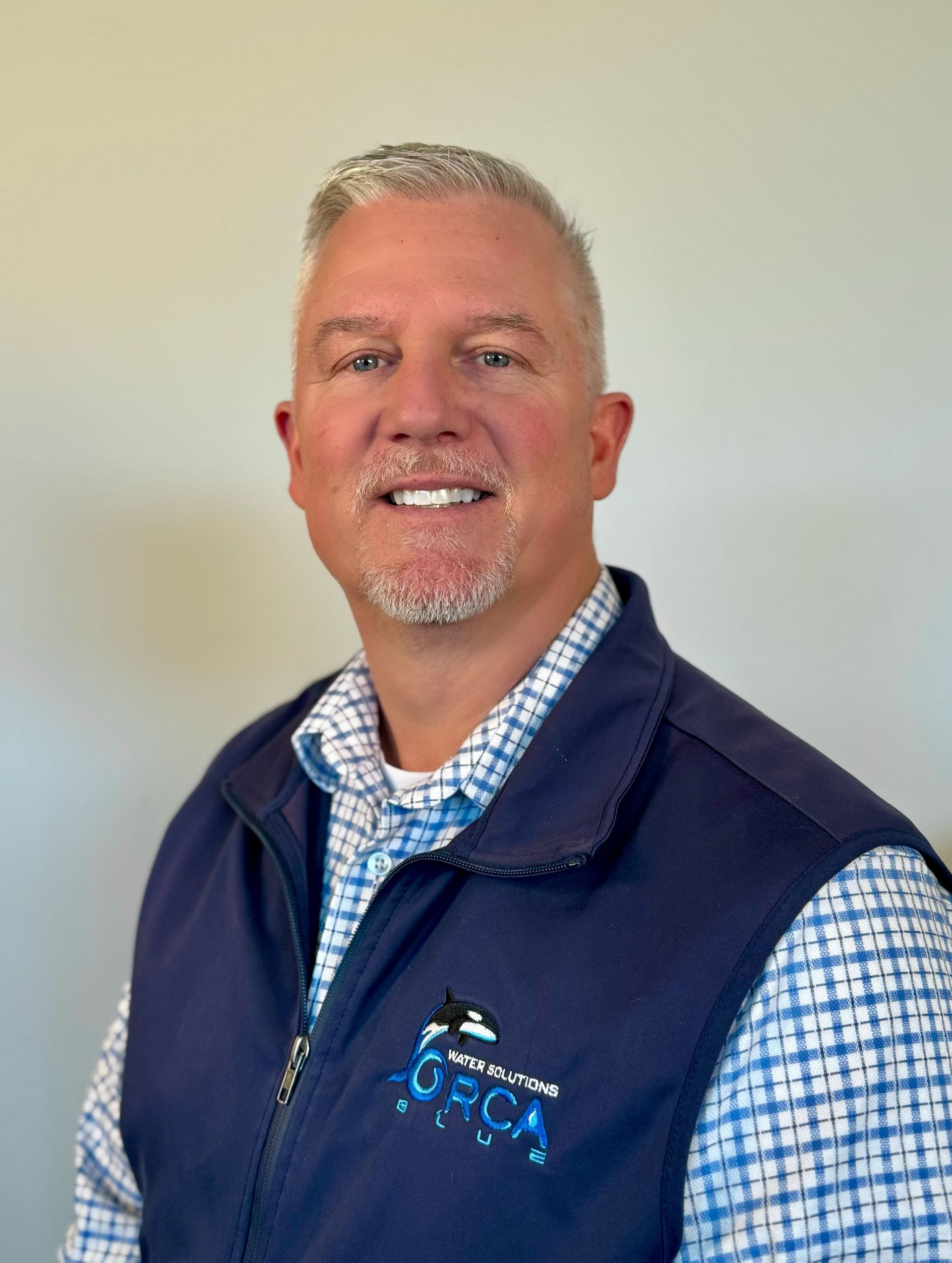 A person with short, light-colored hair and a goatee smiles, wearing a blue patterned shirt under a dark blue vest.