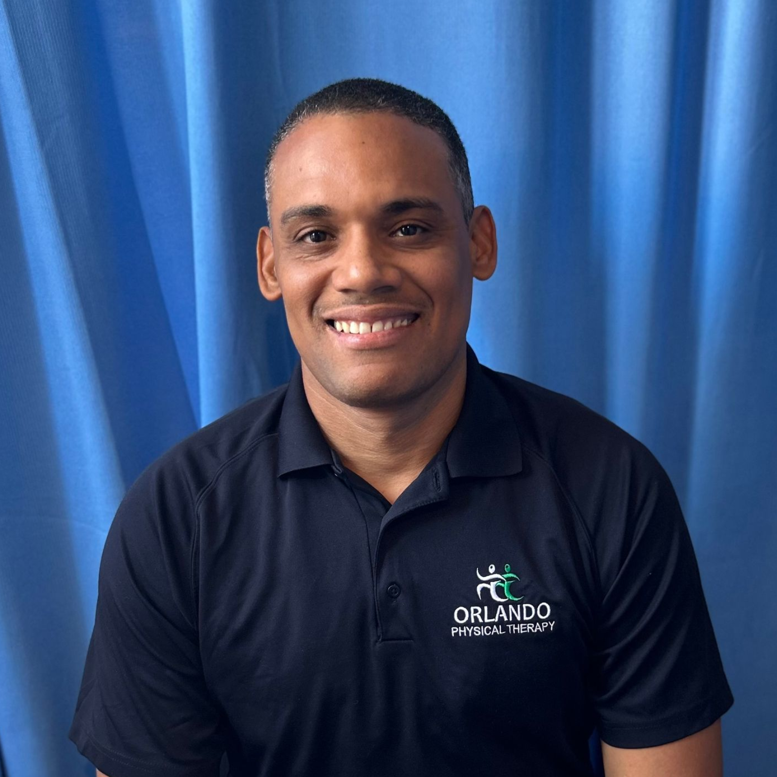 Portrait of a person smiling and wearing a dark collared shirt in front of a blue backdrop.