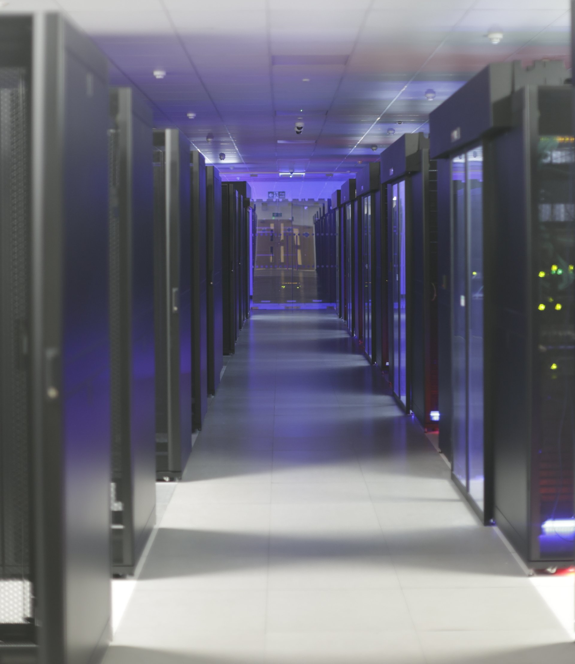 Long hallway in a data center with rows of server racks, lit with blue and white lights.
