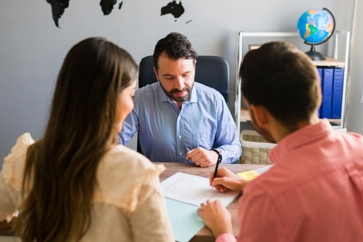 Three people discussing documents at a desk in an office, with a globe and shelves in the background.