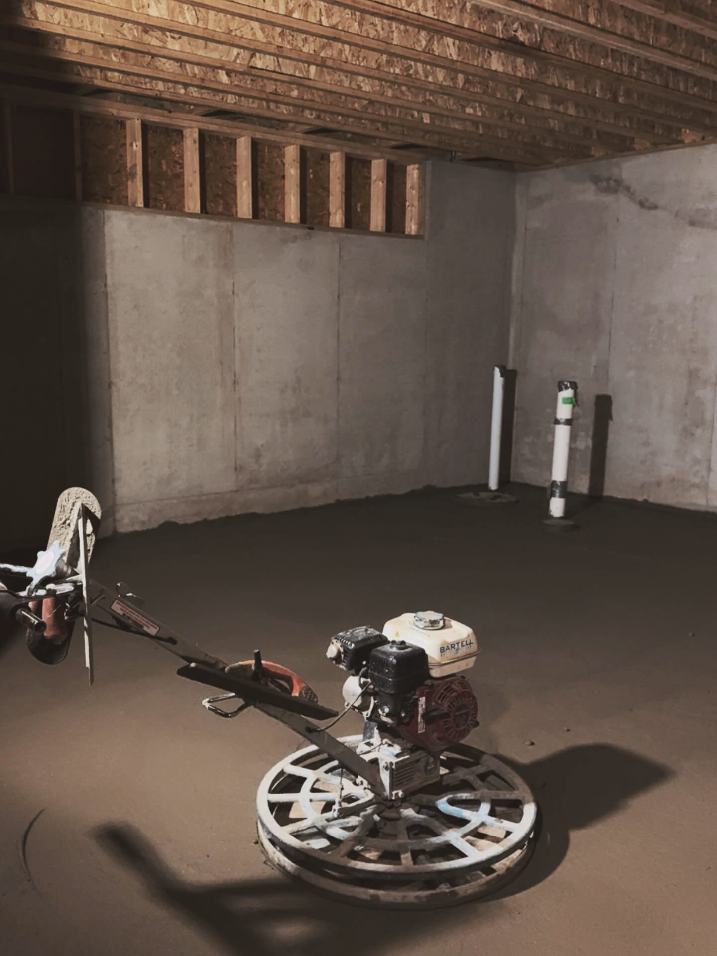Concrete floor being smoothed in a basement with a power trowel, near plumbing and wooden beams.