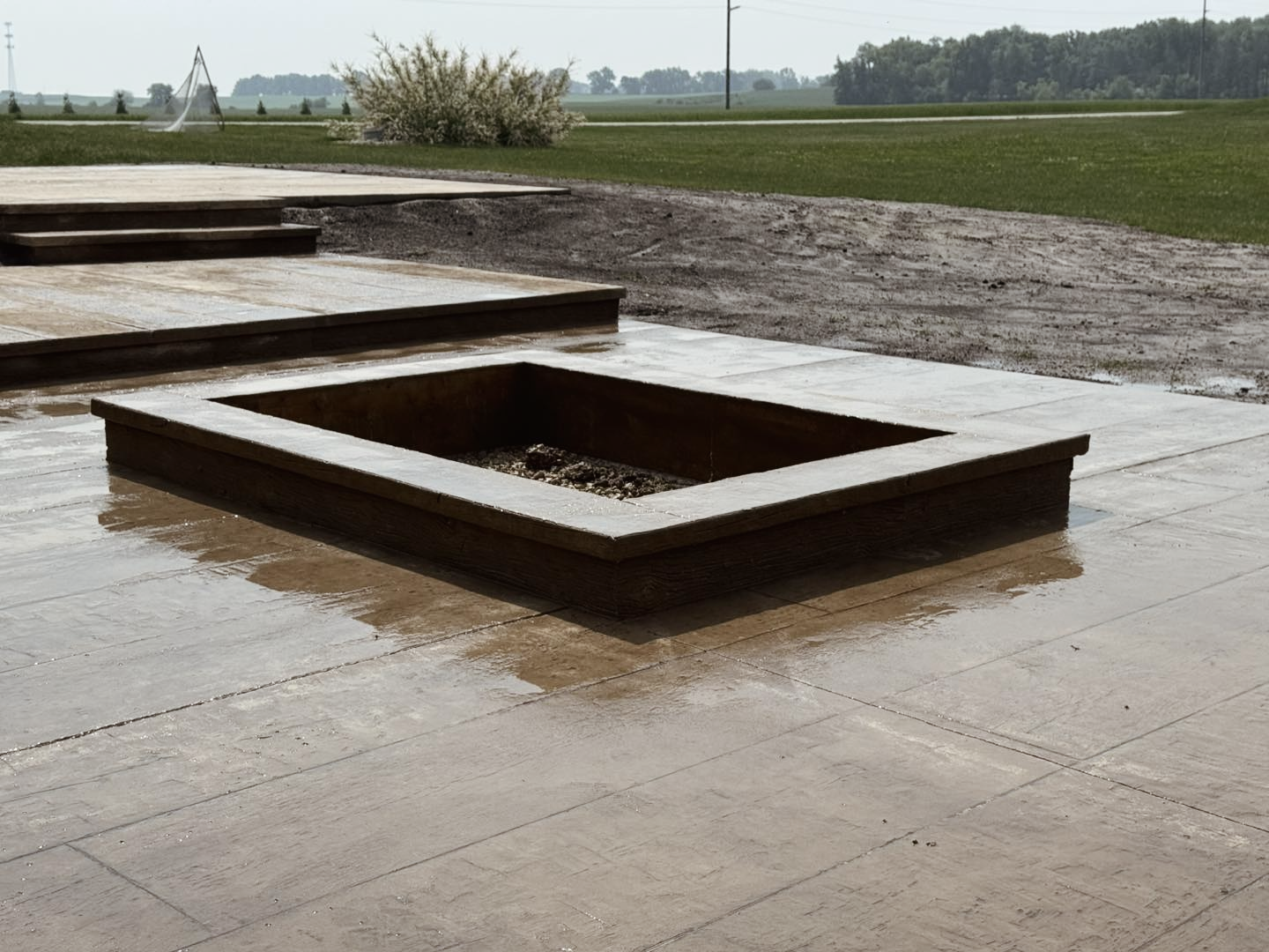 Square fire pit with surrounding patio, wet from rain. Green grass and trees in background.