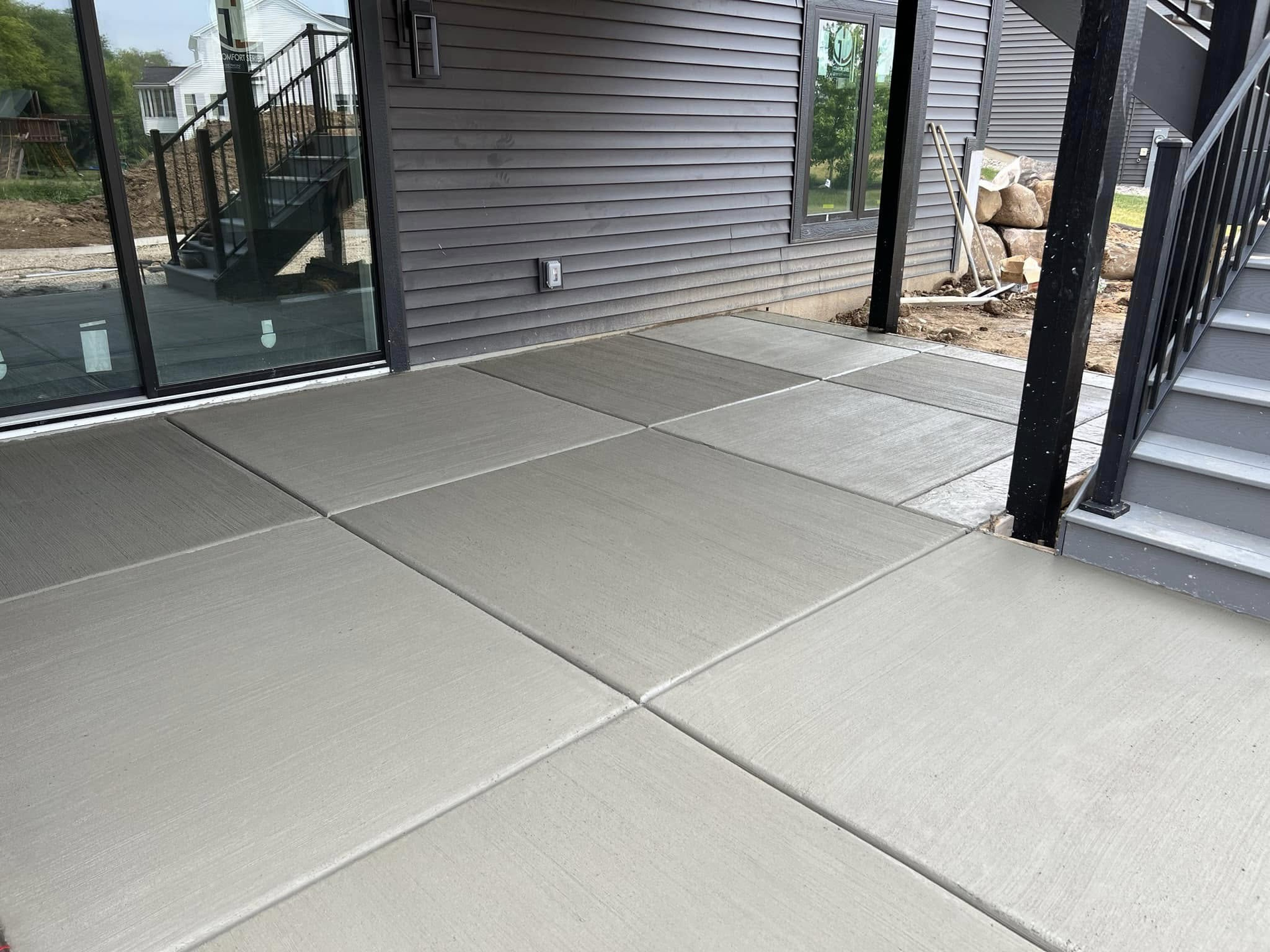 Freshly poured gray concrete patio with square sections next to a dark-sided building and stairs.