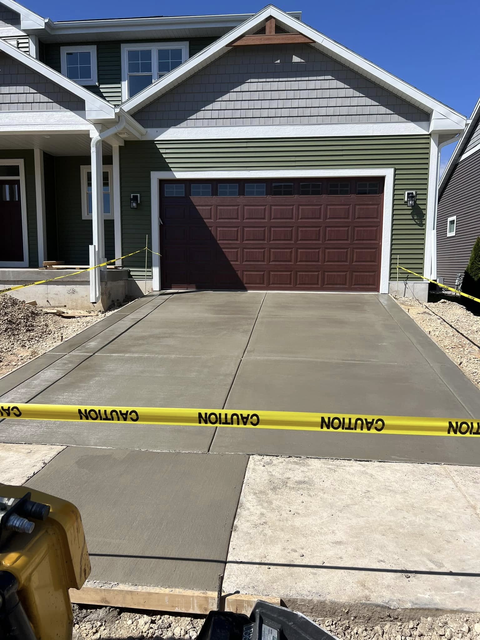 Freshly poured concrete driveway in front of a green house with a brown garage door, yellow caution tape across driveway.