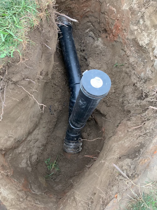 Black pipe in a dirt trench with a capped end, partially buried outdoors.