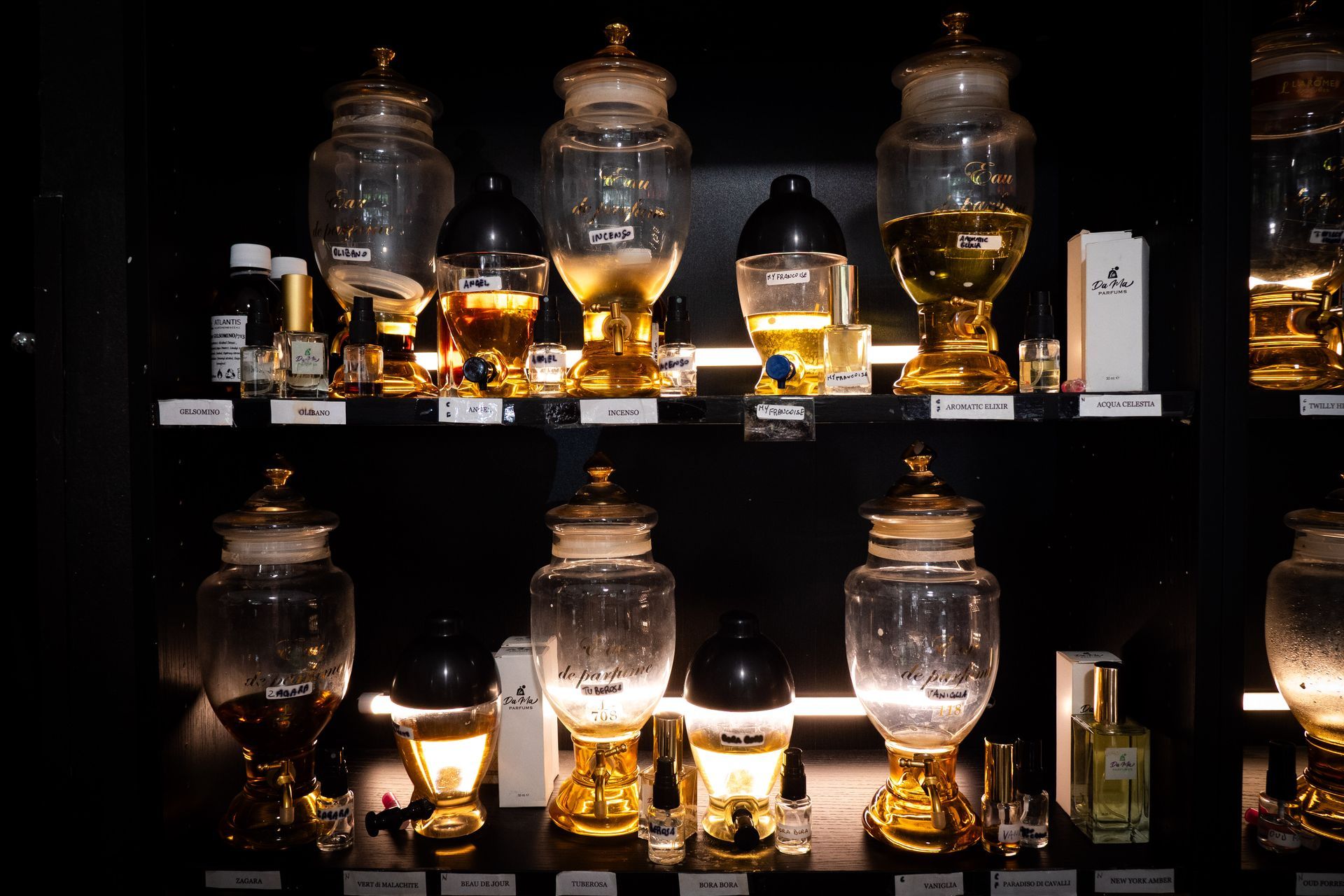 Scaffali pieni di bottiglie di profumo in vetro con liquido dorato, illuminati in un ambiente buio.